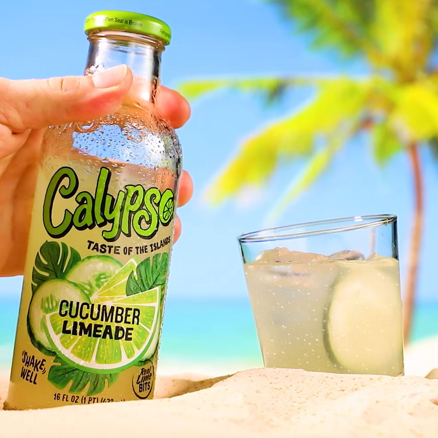 Calypso Cucumber Limeade bottle next to to a cocktail on the beach.