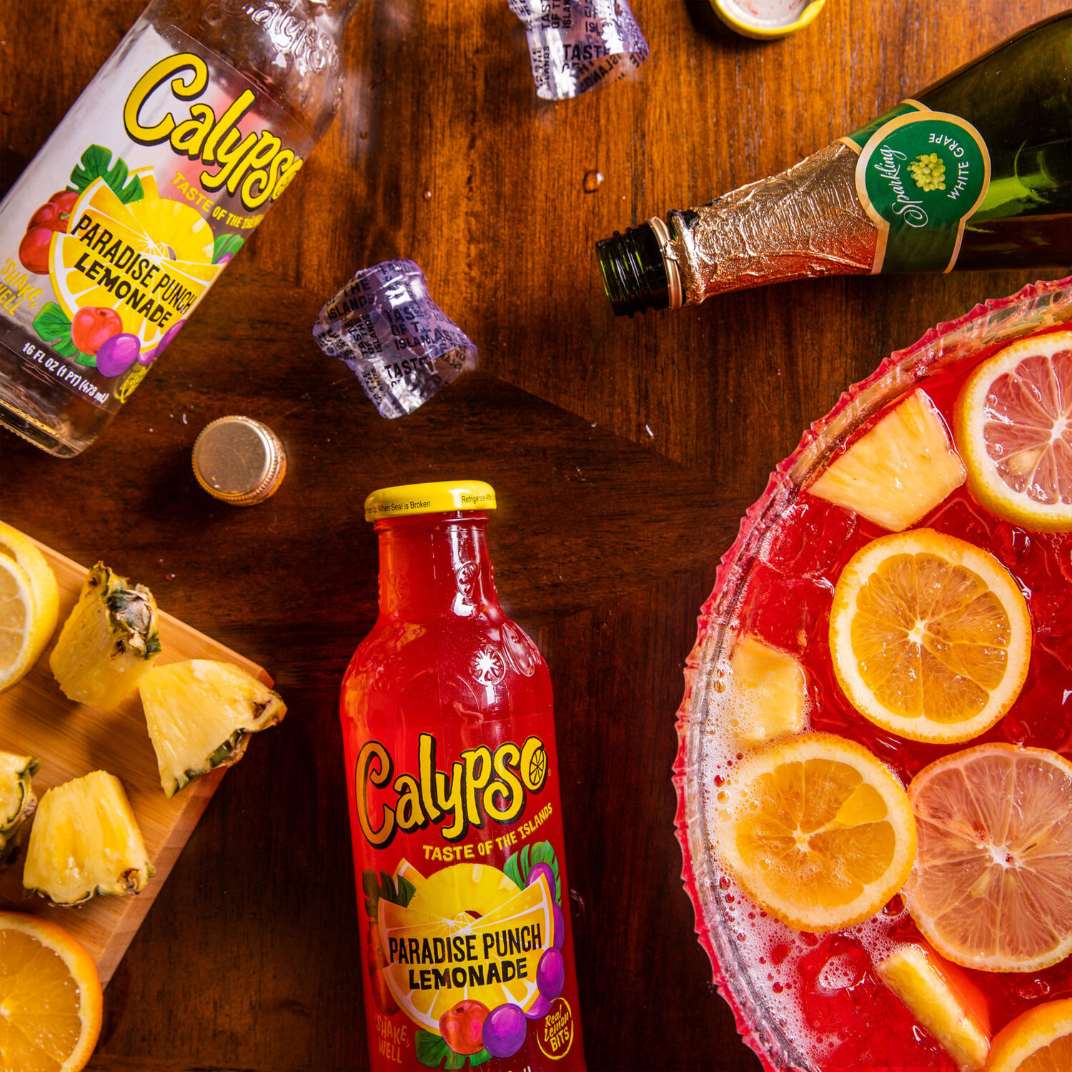 A punch bowl with oranges, pineapples, and lemons next to a bottle of champagne and Calypso Paradise Punch Lemonade.