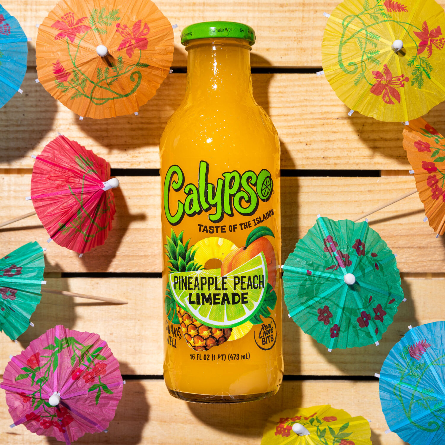 A bottle of Calypso Pineapple Peach Limeade next to umbrellas on a wooden table.