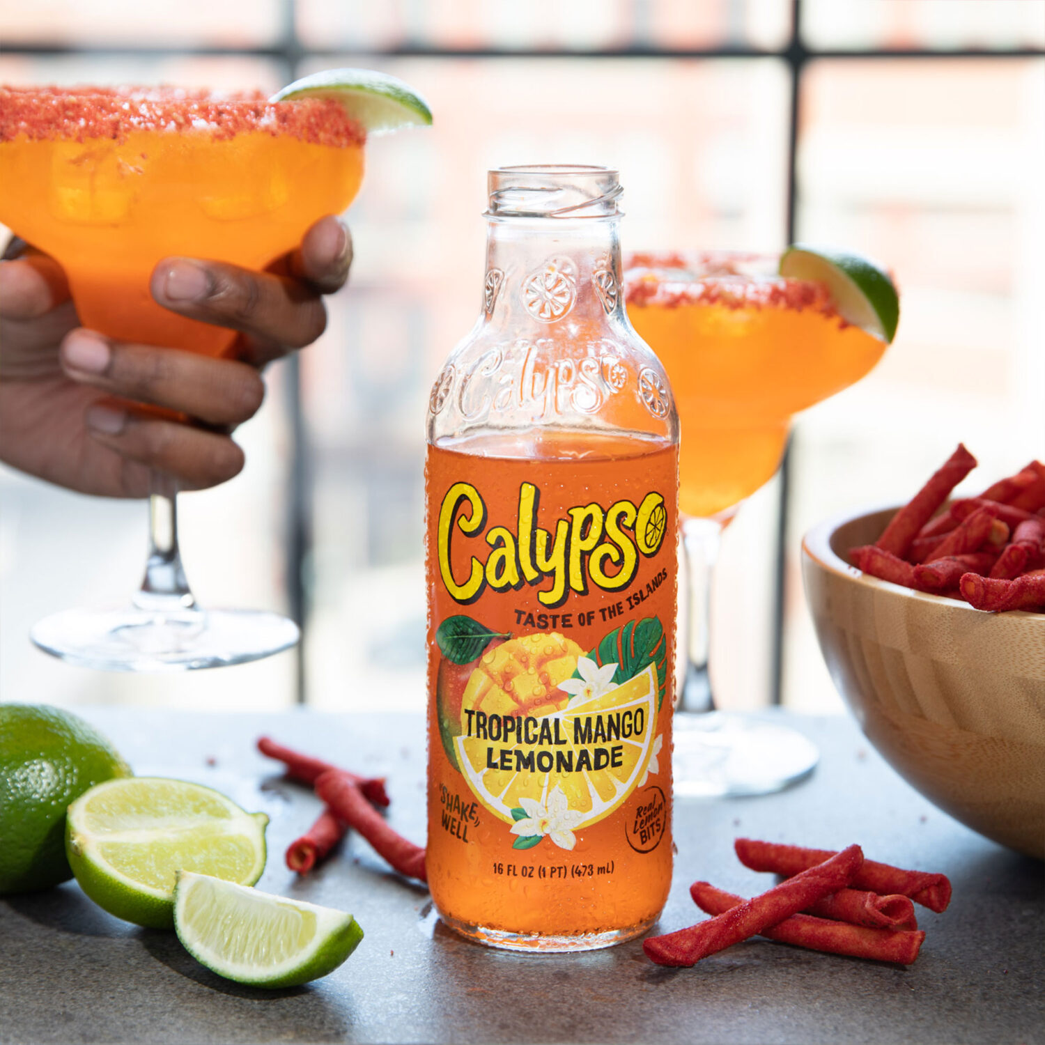 A bottle of Calypso Tropical Mango Lemonade next to a bowl of Takis and a cocktail.